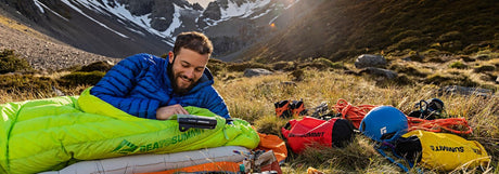 Hiker in a green sleeping bag on an inflatable mat beside their gear, with snowy mountains behind